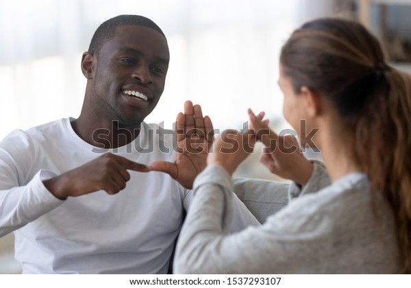 Smiling\
african American man sit on couch show hand gestures talking with\
female friend at home, international disabled hearing impaired\
couple or spouses use sign language\
communicating