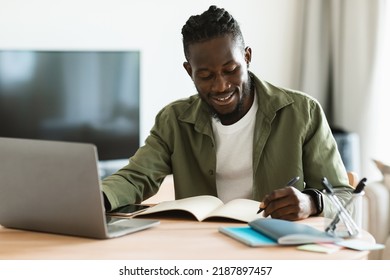 Smiling African American Man Sitting At Desk, Working On Laptop And Taking Notes In Notebook, Black Male Studying Online Or Watching Webinar And Writing Check List