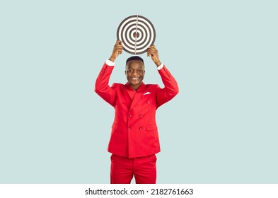 Smiling African American Man In Stylish Red Suit Who Is Holding Target Over His Head On Pastel Light Blue Background. Concept Of Setting Business Goal, Finding Target Audience And Achieving Success.