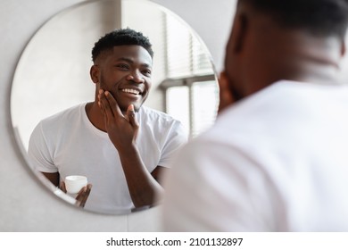 Smiling African American Man Touching Face Applying Moisturizer On Face Holding Moisturizing Cream Jar Looking At His Reflection In Round Mirror Standing In Bathroom At Home. Facial Skincare
