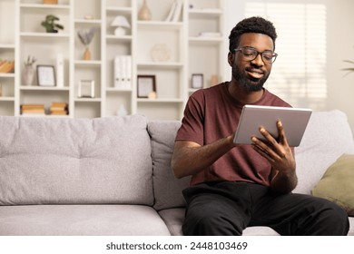 Smiling African American Man Using Tablet On Couch At Home. Casual Lifestyle, Digital Literacy, E-Learning Concept. Joyful Male Engaged In Social Media Or Working Remotely.  - Powered by Shutterstock