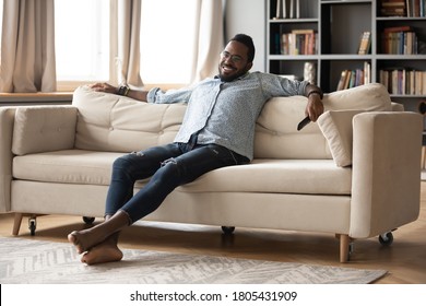 Smiling African American Man Wearing Glasses Relaxing On Cozy Couch In Modern Living Room, Holdin Remote Television Controller In Hand, Positive Young Male Watching Tv Show, Enjoying Lazy Weekend