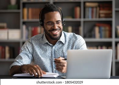 Smiling African American man wearing headphones looking at laptop screen, motivated student writing notes during online lesson, watching webinar, learning language online, sitting at work desk - Powered by Shutterstock
