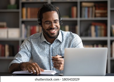 Smiling African American Man Wearing Headphones Looking At Laptop Screen, Motivated Student Writing Notes During Online Lesson, Watching Webinar, Learning Language Online, Sitting At Work Desk