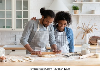 Smiling African American Man And Woman Wearing Aprons Rolling Out Dough, Happy Young Couple Cooking Homemade Pastry Or Pie Together, Using Rolling Pin And Cutters, Standing In Kitchen At Home