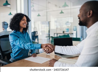 Smiling African American manager sitting at his desk in an office shaking hands with a potential new employee after an interview