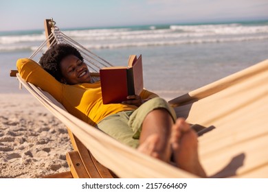 Smiling african american mature woman reading book while lying on hammock against sea in summer.  - Powered by Shutterstock