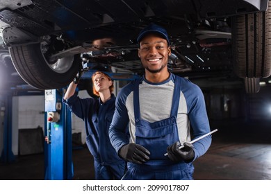 Smiling african american mechanic with digital tablet looking at camera near colleague and car in garage - Powered by Shutterstock