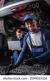 Smiling african american mechanic holding tools near colleague using laptop in car service