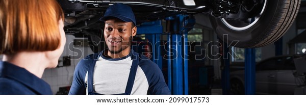 Smiling african american mechanic looking\
at blurred colleague in car service,\
banner