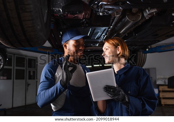 Smiling african\
american mechanic showing ok gesture near colleague with digital\
tablet under car in\
service