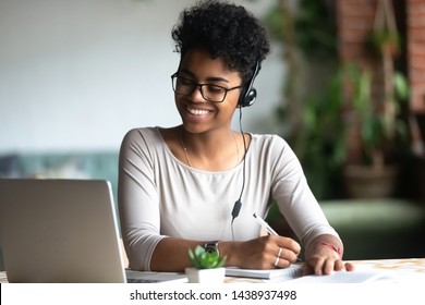 Smiling african American millennial female student in headphones and glasses sit at desk watch webinar making notes, happy biracial young woman in earphones work study using computer write in notebook - Shutterstock ID 1438937498