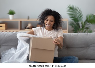 Smiling African American Millennial Girl Sit On Sofa At Home Open Cardboard Delivery Package Shopping Online, Excited Black Young Woman Unpack Unwrap Parcel Buying Good Via Internet Or Web