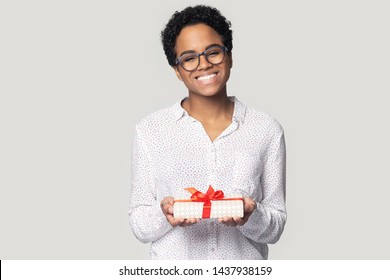 Smiling african American millennial woman in glasses isolated on grey studio background hold wrapped birthday present, happy biracial girl give gift box with red bow greeting with special occasion