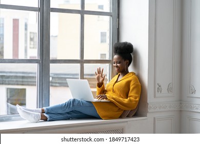 Smiling African American millennial woman with afro hairstyle in yellow cardigan sitting on windowsill, resting, looking at camera webcam, say hello, talking on video chat with her friends on laptop. 