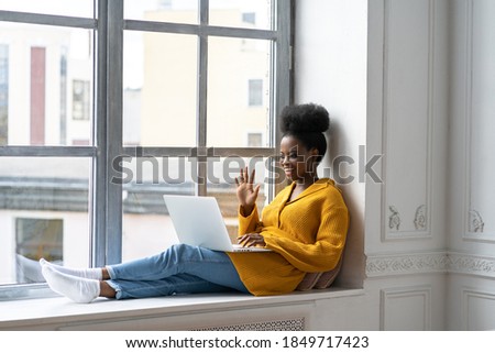 Smiling African American millennial woman with afro hairstyle in yellow cardigan sitting on windowsill, resting, looking at camera webcam, say hello, talking on video chat with her friends on laptop. 