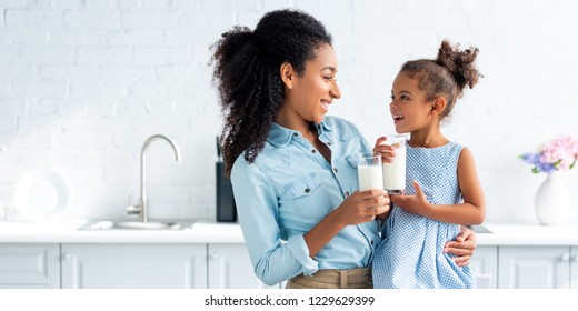 smiling african american mother and daughter holding glasses of milk in kitchen and looking at each other