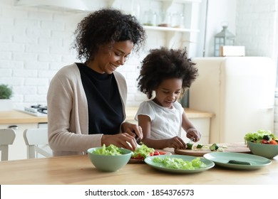 Smiling African American Mother With Daughter Cooking Salad Together, Chopping Fresh Vegetables On Board, Sitting At Wooden Kitchen Table, Caring Mum And Child Preparing Meal, Having Fun