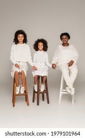 Smiling African American Mother, Daughter And Grandmother Holding Hands And Sitting On Stools On Grey Background