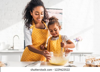 smiling african american mother helping daughter preparing and whisking dough in kitchen