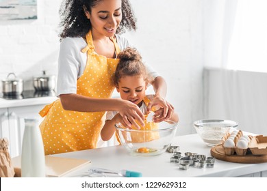 smiling african american mother helping daughter breaking egg for preparing dessert in kitchen - Powered by Shutterstock