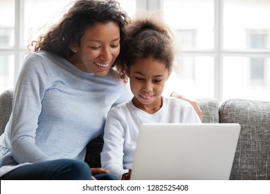 Smiling African American Mother And Kid Daughter Have Fun Shopping Online At Home, Happy Black Mom Teaching Little Mixed Race Child Girl Learn Use Laptop, Watch Cartoon, Make Video Call On Computer