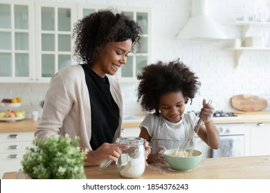 Smiling African American mother with little daughter cooking pancakes, standing at table in kitchen, pretty girl wearing apron using whisk, preparing dough for cookies, family enjoying leisure time - Powered by Shutterstock