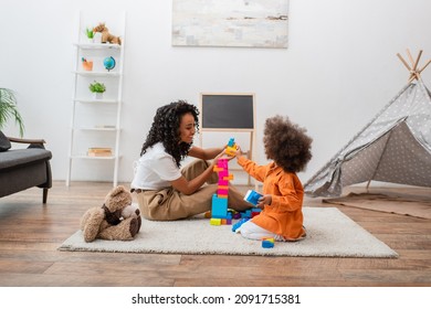 Smiling african american mother playing building blocks with child near teddy bear at home - Powered by Shutterstock