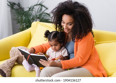 smiling african american mother sitting with toddler daughter and looking at notebook in living room - Shutterstock ID 1904136127