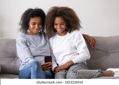 Smiling African American Mother And Teen Daughter Using Smartphone Mobile Apps For Social Media Online Shopping, Happy Black Family Mom And Girl Teenager Having Fun With Phone Sitting On Sofa At Home