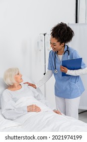 Smiling African American Nurse With Clipboard Touching Senior Patient On Hospital Bed