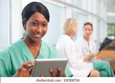Smiling African- American Nurse Holding Tablet With . A Caucasian And Hispanic Doctor In Background, Looking At Scan In Hospital