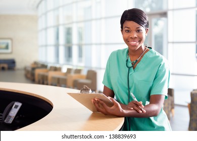 Smiling African American Nurse At Hospital Work Station Lit Brightly With Clipboard And Stethoscope.