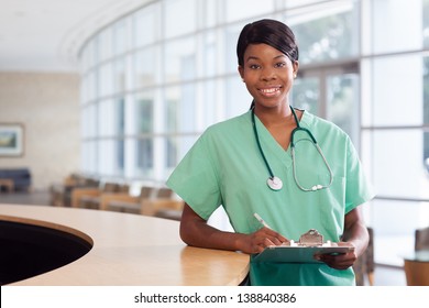 Smiling African American Nurse At Hospital Work Station Lit Brightly With Clipboard And Stethoscope.