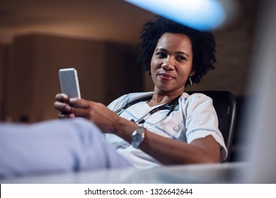 Smiling African American Nurse Relaxing And Typing Text Message On Cell Phone. 