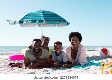 Smiling african american parents and children lying on towels at sandy beach under clear sky. Copy space, unaltered, family, together, parasol, picnic, nature, vacation, enjoyment, relaxing, summer. - Powered by Shutterstock