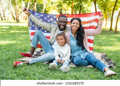 smiling african american parents and daughter holding american flag at picnic in park