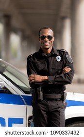 Smiling African American Police Officer With Crossed Arms Leaning Back On Car And Looking At Camera