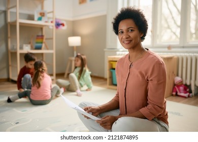 Smiling African American preschool teacher sitting on the floor and looking at camera. There are kids playing in the background. - Shutterstock ID 2121874895