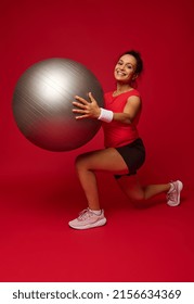 Smiling African American Pretty Woman, Athlete Looking At Camera While Performing Forward Lunges With A Fit Ball Against Red Colored Background With Copy Ad Space