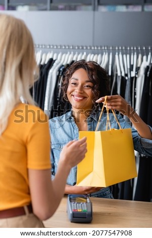 smiling african american saleswoman giving yellow paper bag with purchase to blonde client in boutique