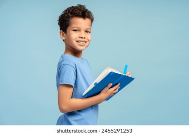 Smiling African American schoolboy holding exercise book while making notes, looking at camera, isolated on blue background. Back to school concept - Powered by Shutterstock