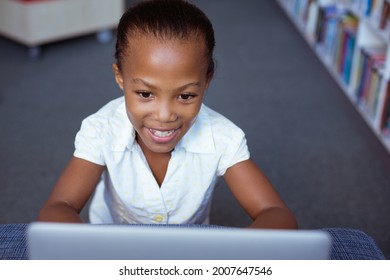 Smiling African American Schoolgirl At Desk In School Library Using Laptop. Childhood, Technology And Education At Elementary School.