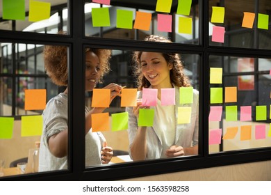 Smiling african american scrum mater working with happy caucasian pleasant colleague, improving IT business project workflow on glass window kanban agile brainstorming board at modern office.