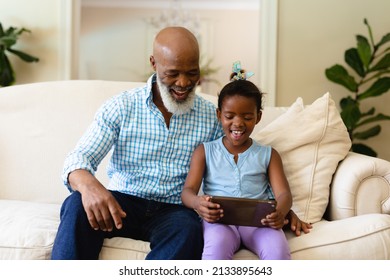 Smiling African American Senior Man With Granddaughter Using Digital Tablet While Sitting On Sofa. Unaltered, Wireless Technology, Family, Lifestyle, Leisure Activity And Domestic Life Concept.