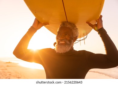 Smiling african american senior man carrying surfboard on head at beach against clear sky at sunset. copy space, sunlight, nature, unaltered, retirement, aquatic sport, holiday and active lifestyle.