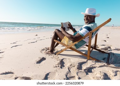 Smiling african american senior man wearing hat reading book on deckchair at beach under clear sky. Copy space, hobbies, unaltered, lifestyle, vacation, retirement, relaxation and nature concept. - Powered by Shutterstock