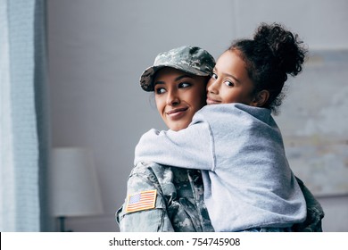 smiling african american soldier in military uniform holding daughter on arms at home