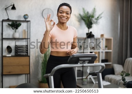 Smiling african american sporty woman in pink t-shirt leaned her hands on treadmill while working out in morning at living room at home, looking at camera showing sign ok.
