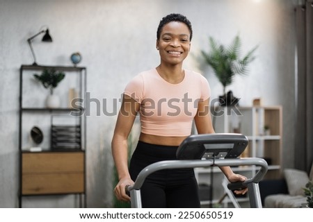 Smiling african american sporty woman in pink t-shirt leaned her hands on treadmill while working out in morning at living room at home, looking at camera.